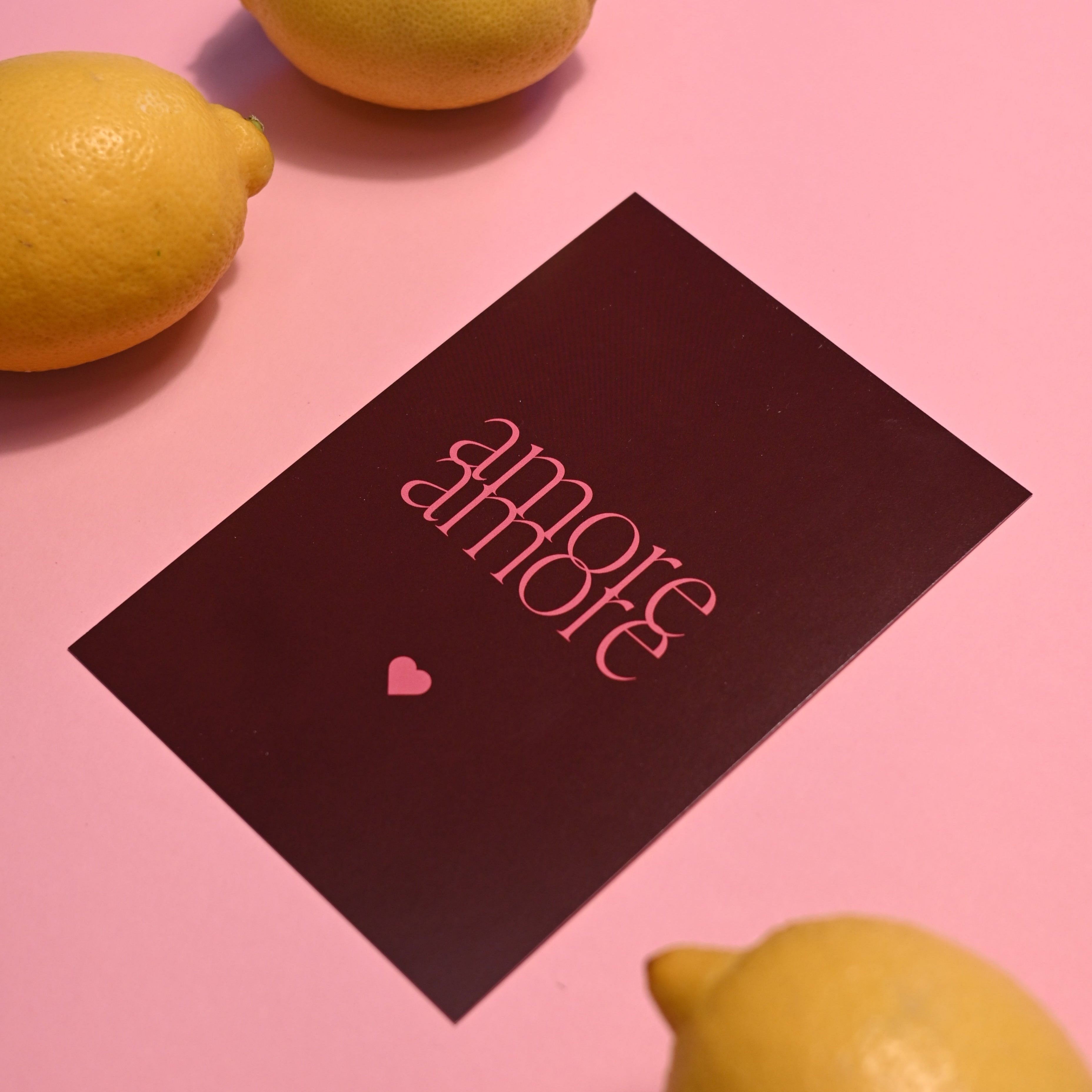 Stilvolle "Amore Amore"-Karte mit pinkem Text, arrangiert auf rosafarbenem Hintergrund, umgeben von gelben Zitronen, passend für elegante Designs.