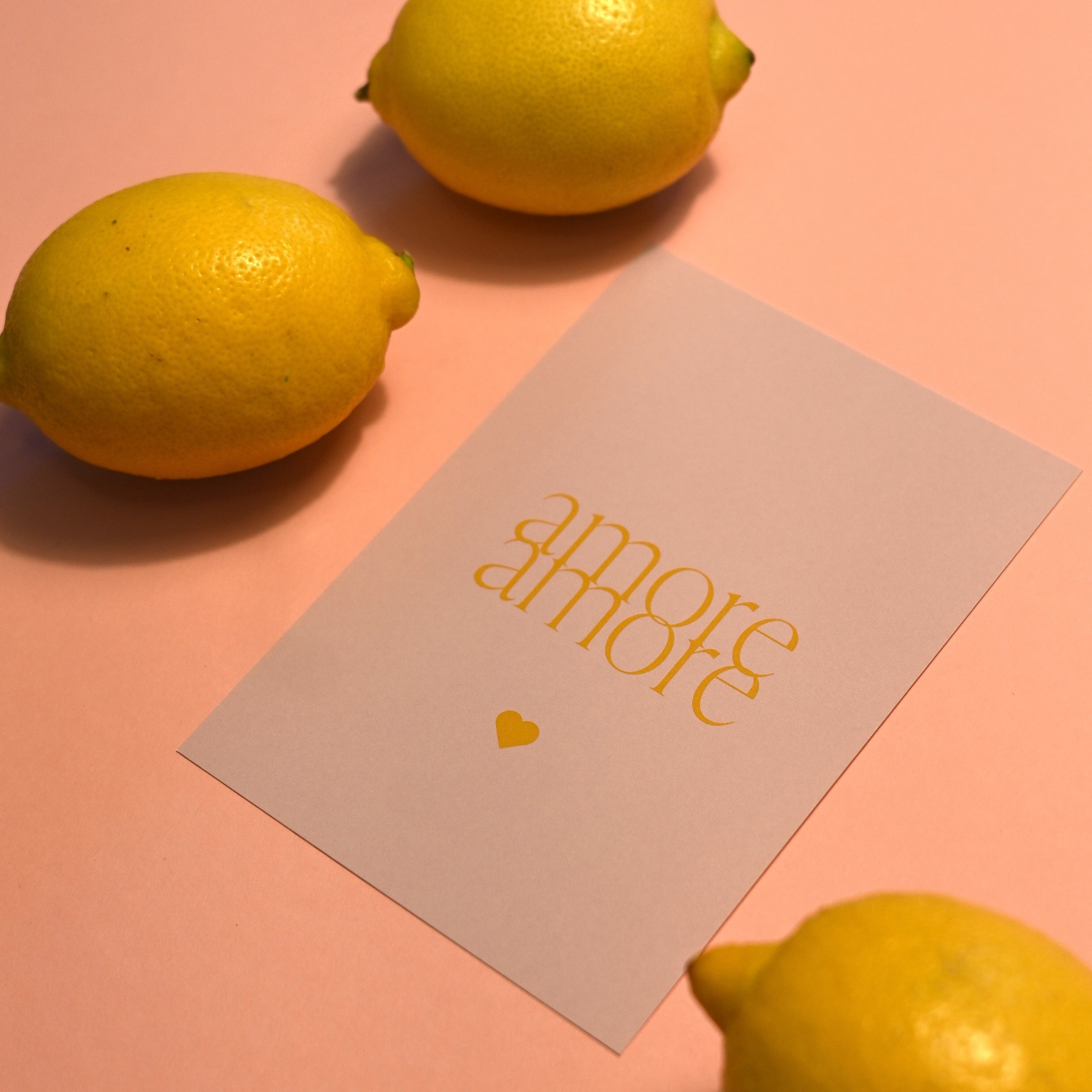 Stilvolle "Amore Amore" Postkarte mit gelbem Text, arrangiert auf rosafarbenem Hintergrund, umgeben von gelben Zitronen, passend für elegante Designs. bei kathis papeterie